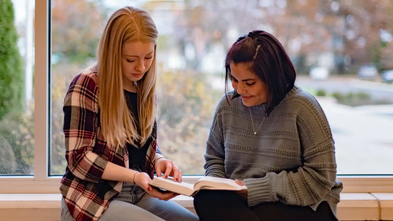Two students sitting by a window, reading the Bible.